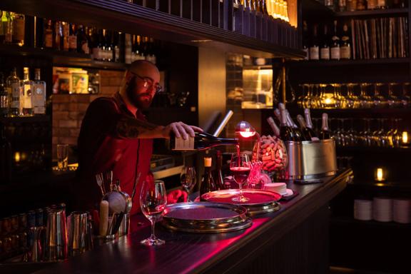 A bartender pours a glass of red wine in a dark cozy wine bar.