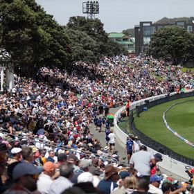 Crowds of people sit on the bank at the Cello Basin Reserve watching a cricket game.