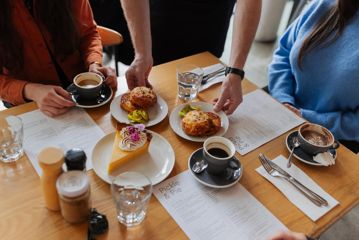 Two cheese scones and a slice of lemon meringue pie are placed on a table alongside a couple of coffees and a hot chocolate.