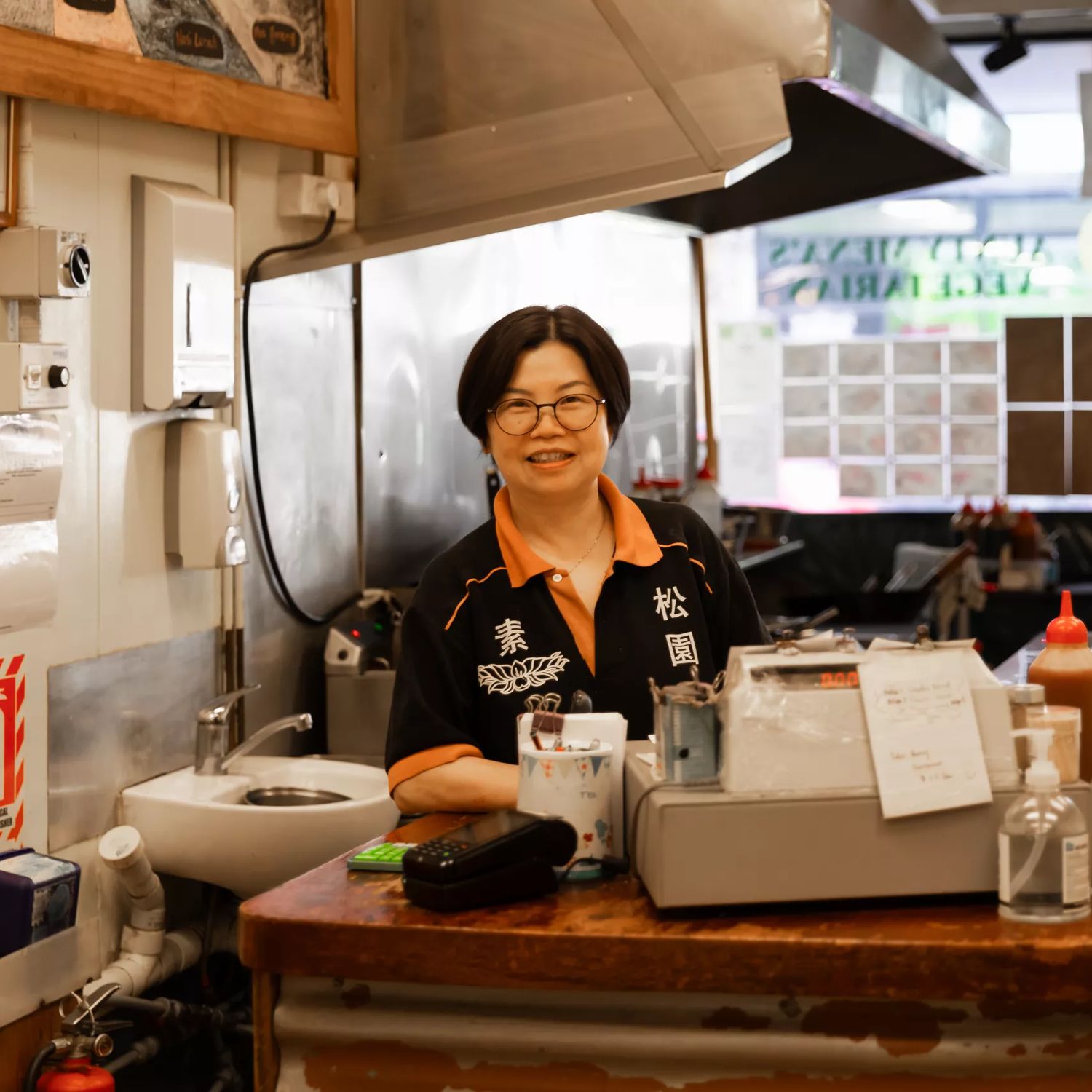 A person standing behind the counter in Aunty Mena's, with dishes, condiments, and a cash register visible.