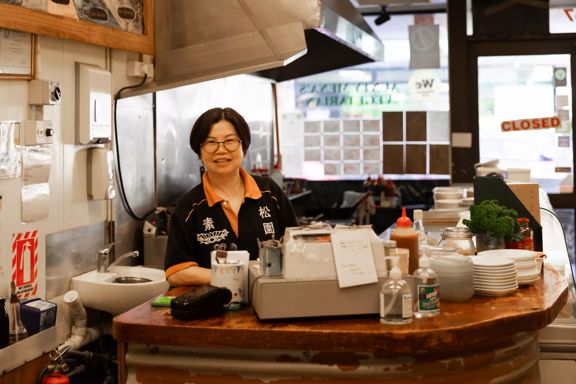 A person standing behind the counter in Aunty Mena's, with dishes, condiments, and a cash register visible.