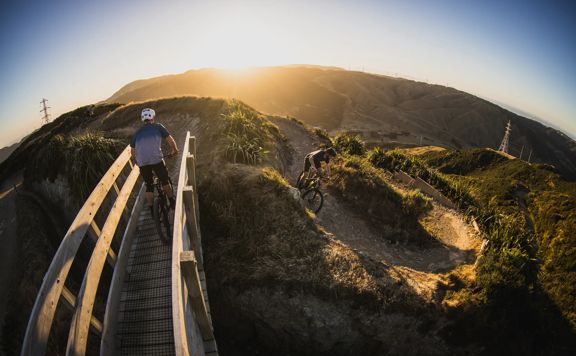 At sunrise, two mountain bikers ride along the winding path at Mākara Peak Mountain Bike Park.
