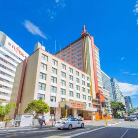 The exterior of the James Cook Hotel Grand Chancellor on a sunny day. It's a beige, rectangular building and there is a white taxi stopped at a red light on the street in front of the hotel. 