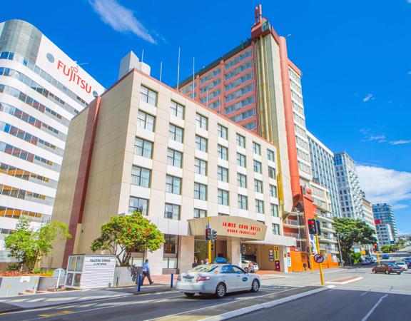 The exterior of the James Cook Hotel Grand Chancellor on a sunny day. It's a beige, rectangular building and there is a white taxi stopped at a red light on the street in front of the hotel. 