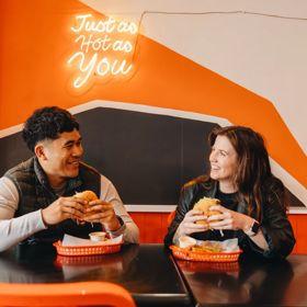 Two people in a vibrant restaurant setting, holding burgers smiling at each other