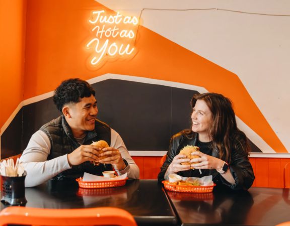 Two people in a vibrant restaurant setting, holding burgers smiling at each other