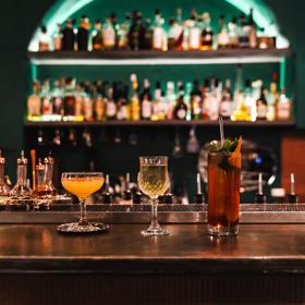 Three cocktails sit in line on a wooden bar. Bottles of alcohol sit on shelves on the wall behind.