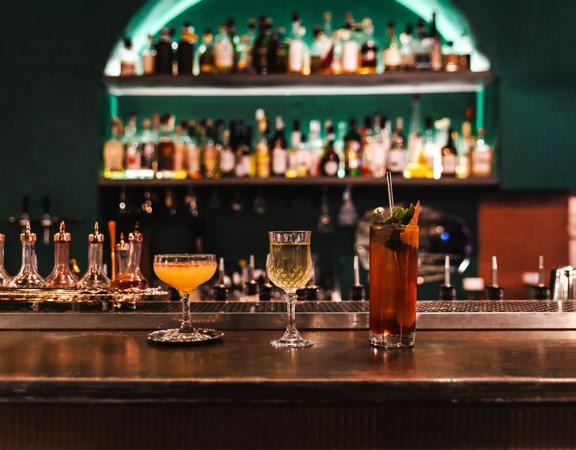 Three cocktails sit in line on a wooden bar. Bottles of alcohol sit on shelves on the wall behind.