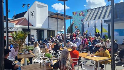 Image of a very busy forecourt outside Toi Mahara. Multiple people are sitting and standing filling the space. The gallery building is in the background.