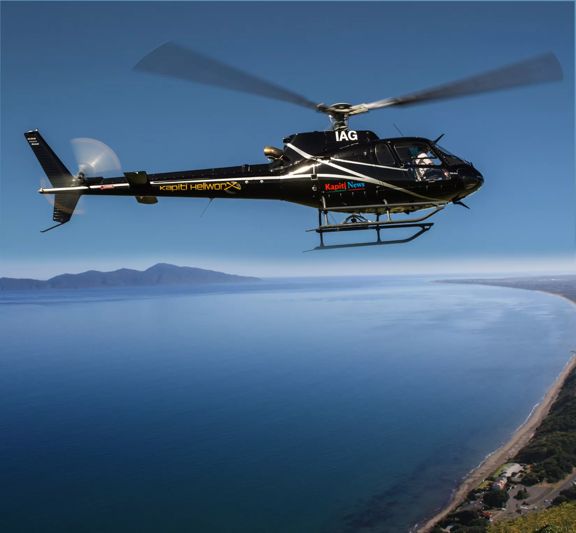 A black helicopter from Kāpiti Heliworx, flying over the Kāpiti Coast on a sunny day.