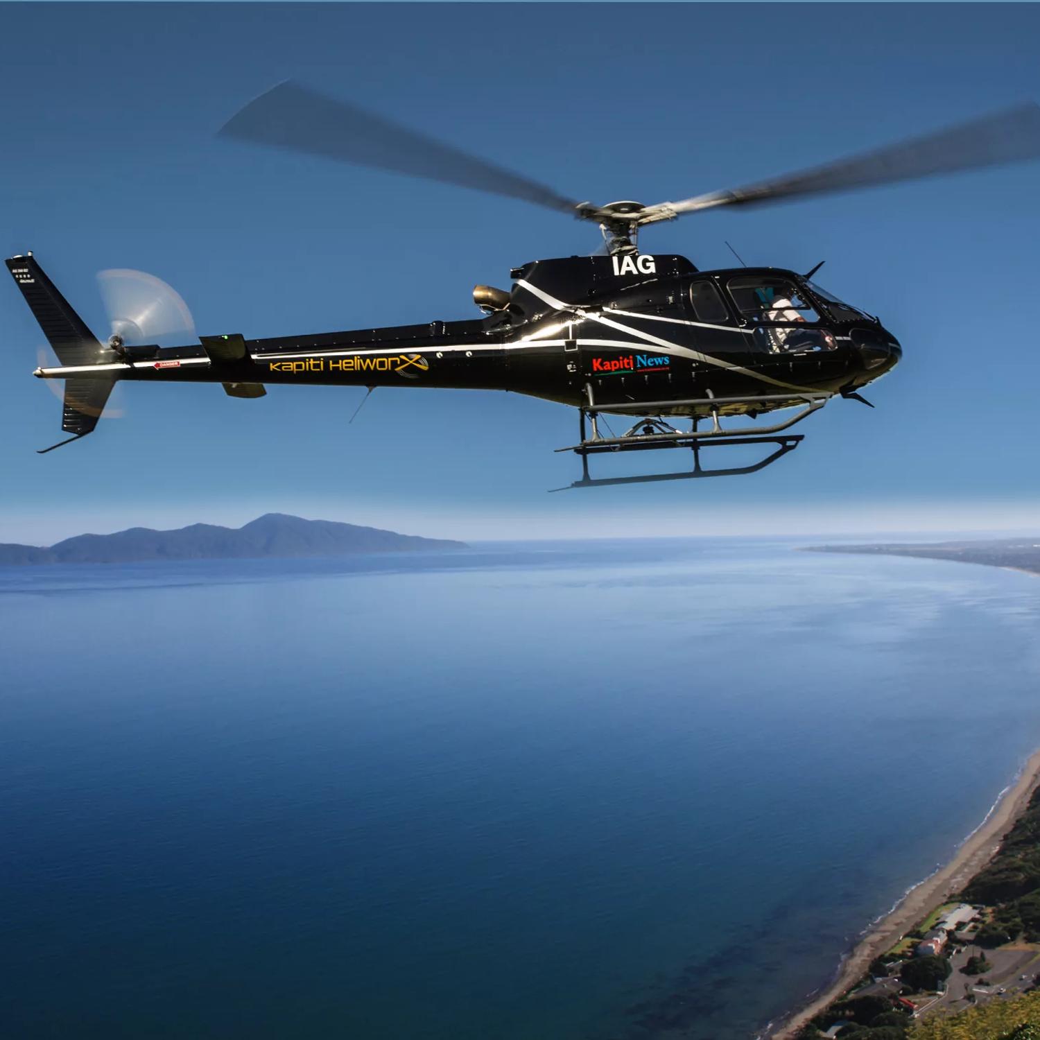 A black helicopter from Kāpiti Heliworx, flying over the Kāpiti Coast on a sunny day.