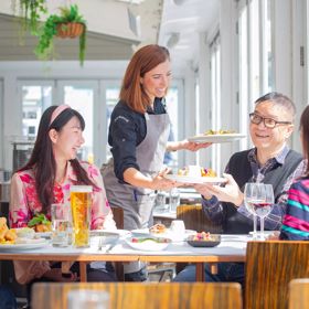 A waiter serves a plate of food to a customer inside Dockside on Queens Wharf. Four people sitting in the sun smile and enjoy their food.
