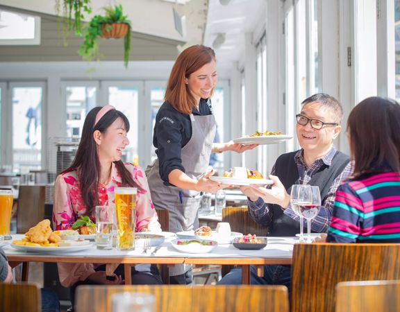 A waiter serves a plate of food to a customer inside Dockside on Queens Wharf. Four people sitting in the sun smile and enjoy their food.