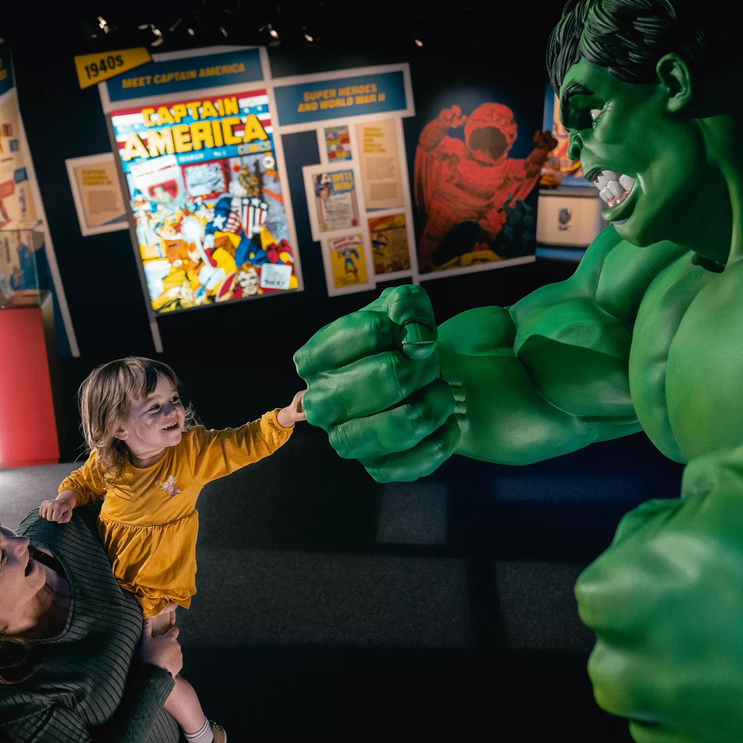 A child in a yellow dress is being held up by a person inside Marvel: Earth's Mightiest Exhibition. Both are smiling while the child fist bumps a statue of the Incredible Hulk. 