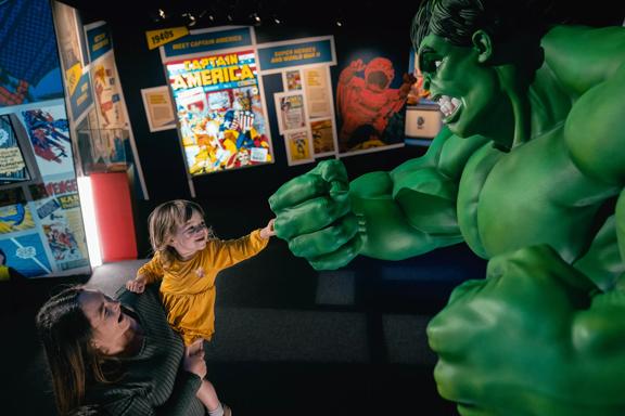 A child in a yellow dress is being held up by a person inside Marvel: Earth's Mightiest Exhibition. Both are smiling while the child fist bumps a statue of the Incredible Hulk. 