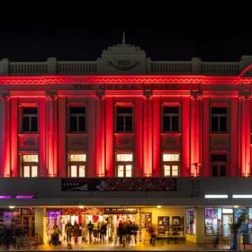 The exterior of The Opera House in Wellington at night. People are walking in and out of the front doors and the top two stories of the building are lit up in red-coloured spotlights.