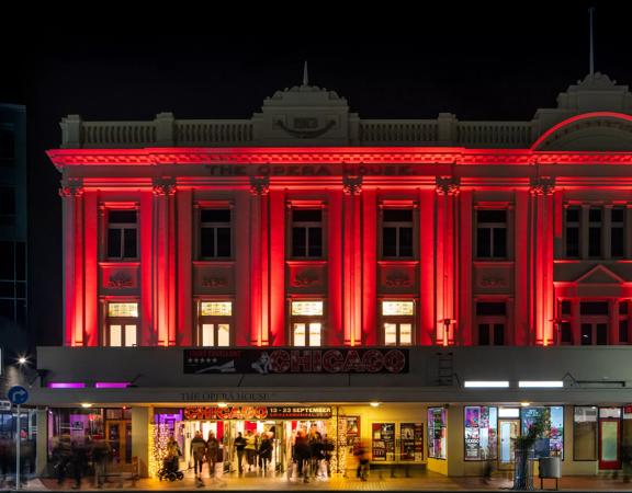 The exterior of The Opera House in Wellington at night. People are walking in and out of the front doors and the top two stories of the building are lit up in red-coloured spotlights.