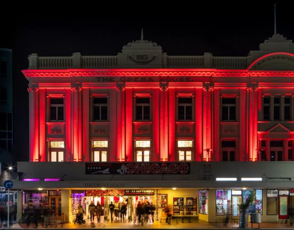 The exterior of The Opera House in Wellington at night. People are walking in and out of the front doors and the top two stories of the building are lit up in red-coloured spotlights.