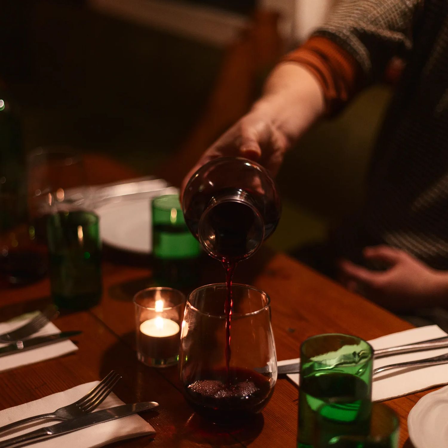 A person pours red wine into a glass at a warmly lit dinner table set with plates, cutlery, green drinking glasses, and candlelight.
