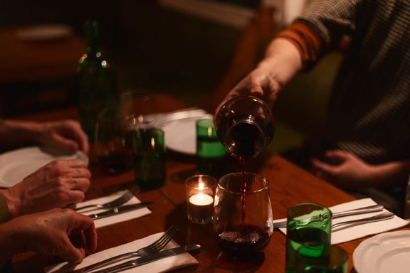 A person pours red wine into a glass at a warmly lit dinner table set with plates, cutlery, green drinking glasses, and candlelight.