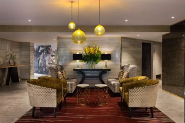 A lounge area in the Mövenpick Hotel lobby. Four small green armchairs and two large grey-patterned armchairs sit on a crimson and gold striped rug. Two small, circular coffee tables sit in the middle.