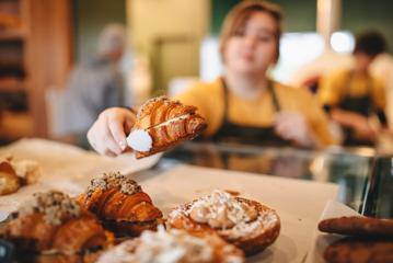 A croissant is picked up with a pair of tongs by a baker wearing a yellow teeshirt and dark grey apron.