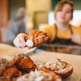 A croissant is picked up with a pair of tongs by a baker wearing a yellow teeshirt and dark grey apron.