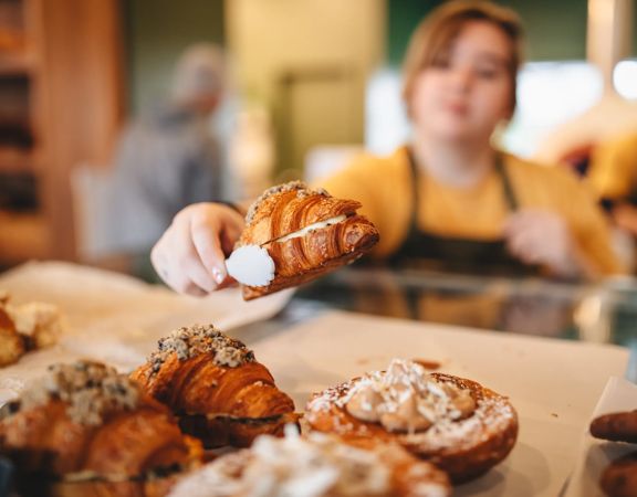 A croissant is picked up with a pair of tongs by a baker wearing a yellow teeshirt and dark grey apron.