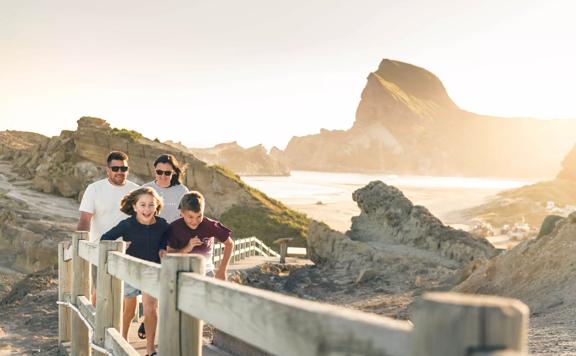 A family walks along a wooden path near Castlepoint, rocks in the background are lit up by the glaring sun.