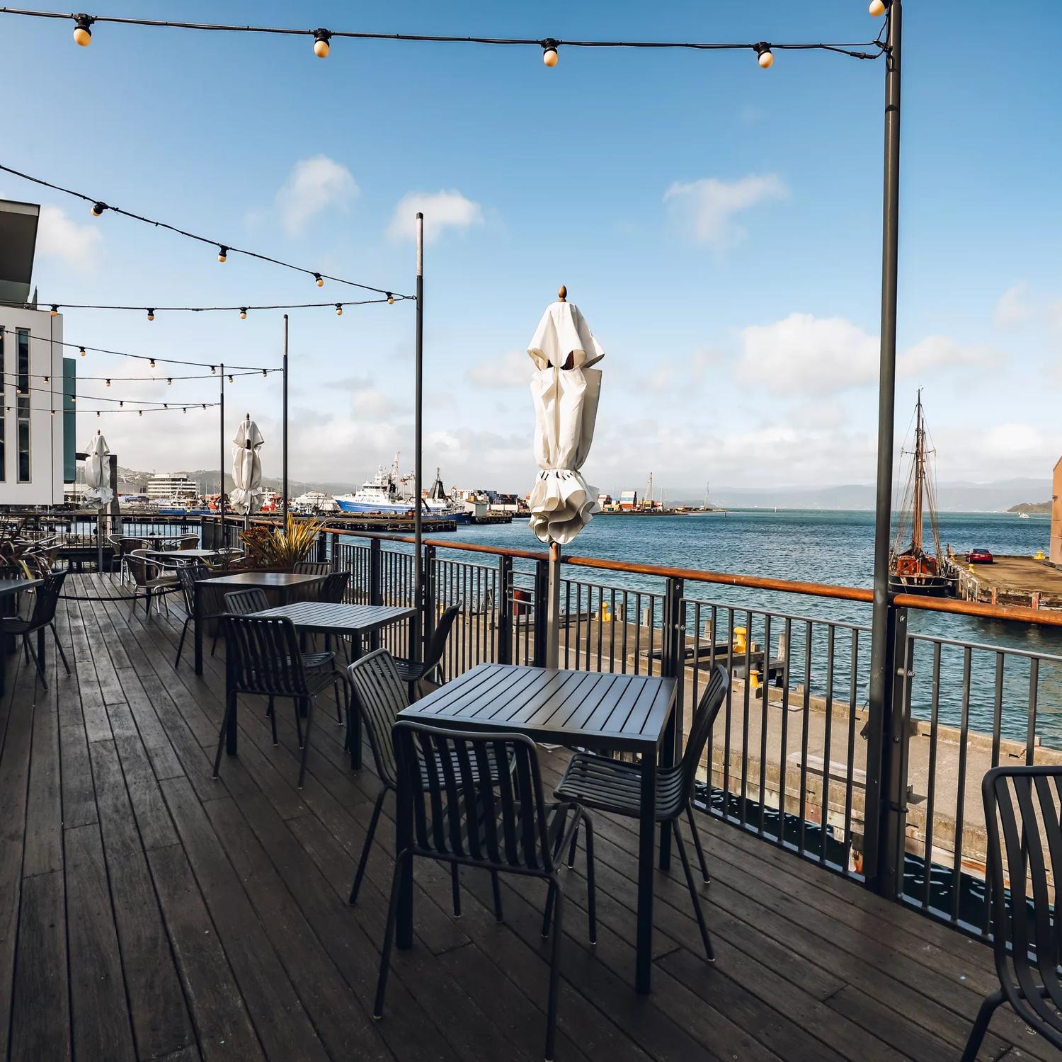 A waterfront terrace with black iron tables and chairs, patio string lights hanging above and a view of the bay and blue sky dotted with a few clouds.