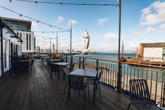 A waterfront terrace with black iron tables and chairs, patio string lights hanging above and a view of the bay and blue sky dotted with a few clouds.