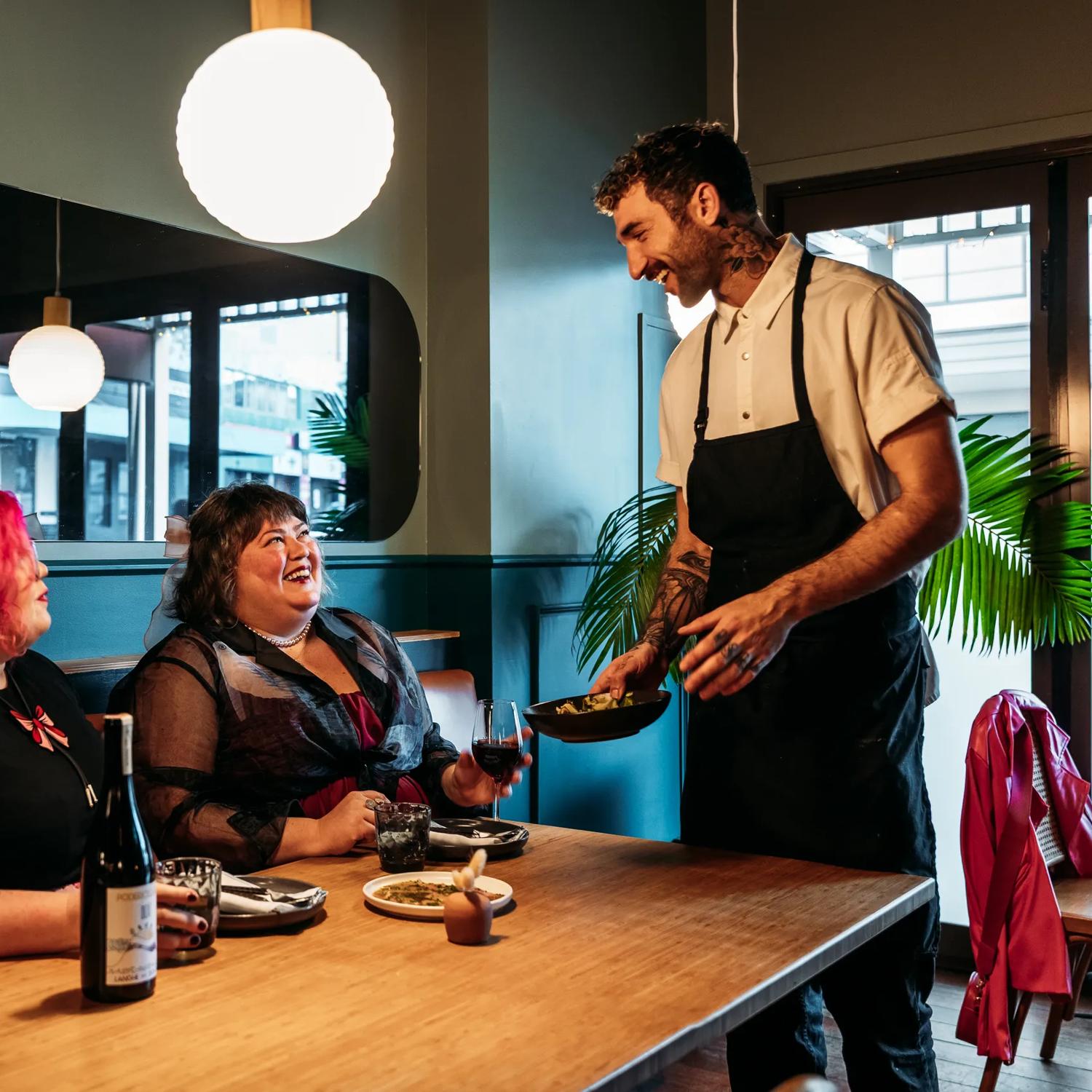 Two people sharing plates of food and a bottle of wine in Koji. There is a waiter handing them a plate and smiling at them. The background is moody, with lantern lights and leather and blue velvet bench seats.