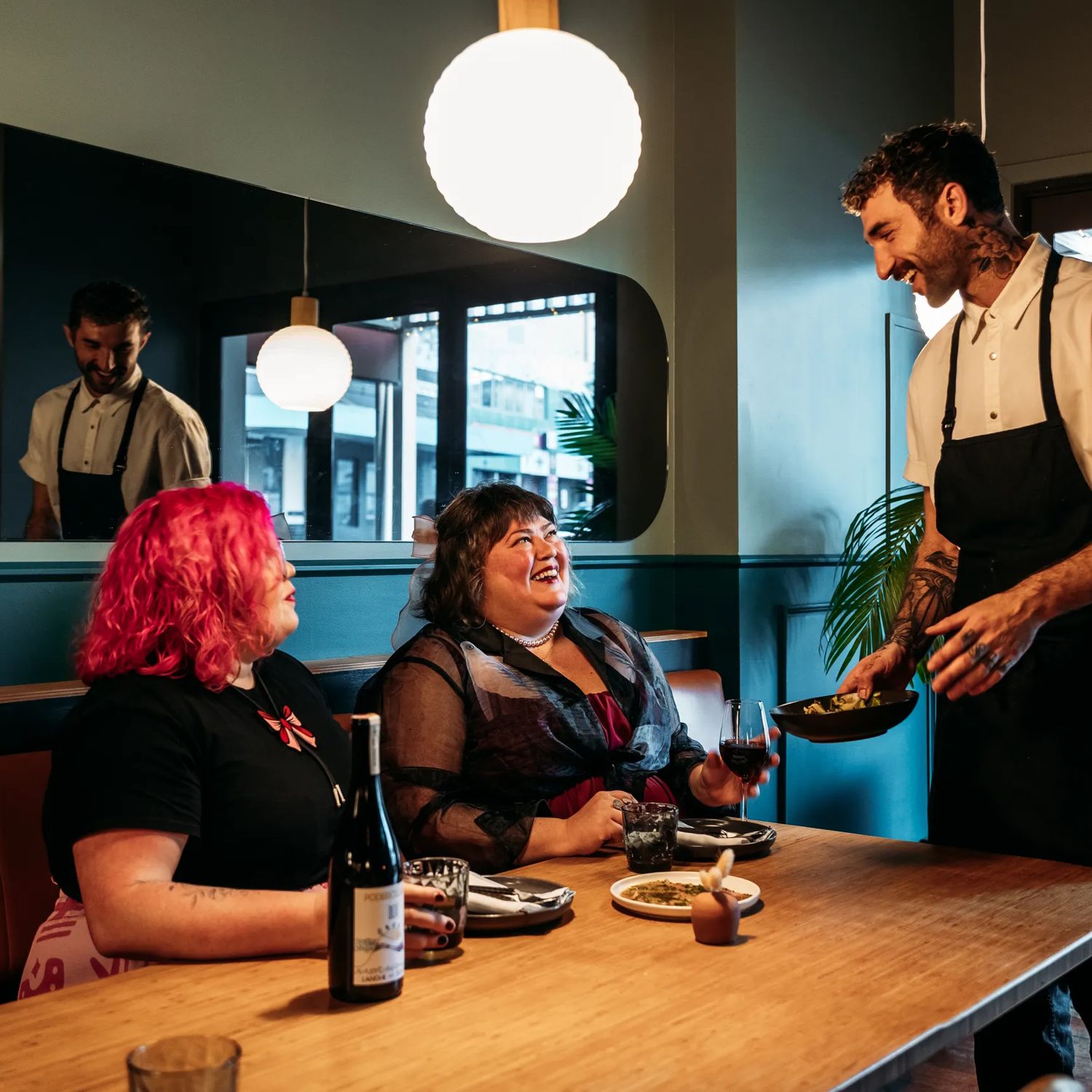 Two people sharing plates of food and a bottle of wine in Koji. There is a waiter handing them a plate and smiling at them. The background is moody, with lantern lights and leather and blue velvet bench seats.