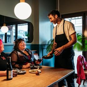 Two people sharing plates of food and a bottle of wine in Koji. There is a waiter handing them a plate and smiling at them. The background is moody, with lantern lights and leather and blue velvet bench seats.