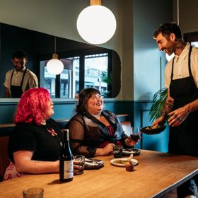 Two people sharing plates of food and a bottle of wine in Koji. There is a waiter handing them a plate and smiling at them. The background is moody, with lantern lights and leather and blue velvet bench seats.