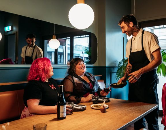 Two people sharing plates of food and a bottle of wine in Koji. There is a waiter handing them a plate and smiling at them. The background is moody, with lantern lights and leather and blue velvet bench seats.