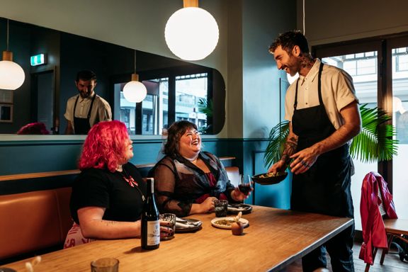 Two people sharing plates of food and a bottle of wine in Koji. There is a waiter handing them a plate and smiling at them. The background is moody, with lantern lights and leather and blue velvet bench seats.