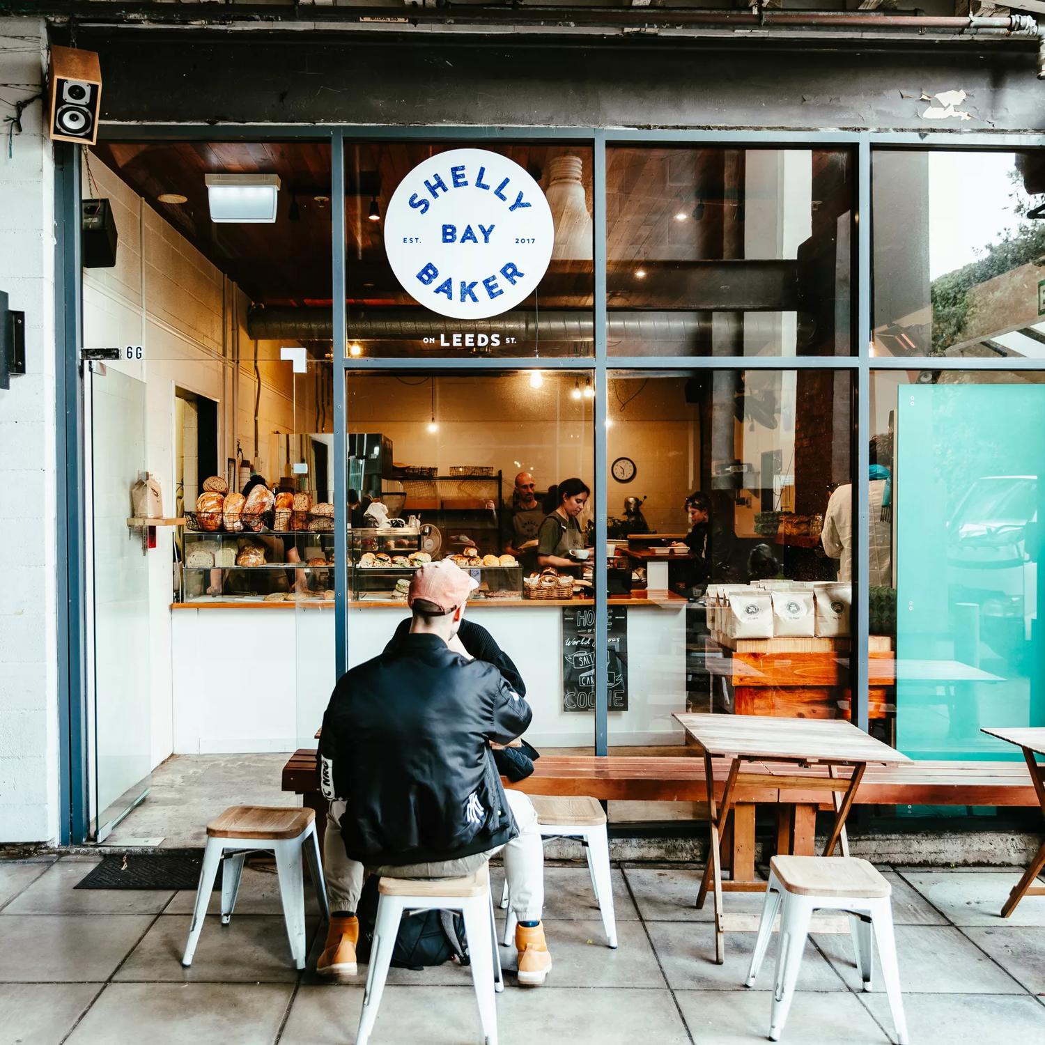 The front window of Shelly Bay Bakery located on Leeds Street in Te Aro in Wellington. Two people are seated at one of the three small wooden tables. 