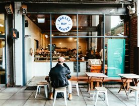 The front window of Shelly Bay Bakery located on Leeds Street in Te Aro in Wellington. Two people are seated at one of the three small wooden tables.