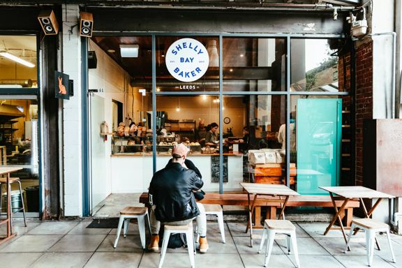 The front window of Shelly Bay Bakery located on Leeds Street in Te Aro in Wellington. Two people are seated at one of the three small wooden tables.