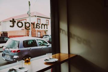 The view from the front window of Margot, a restaurant in Newtown, Wellington, New Zealand. The restaurant's name is printed on the window and two place settings are on the wooden counter.