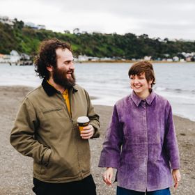 Two people walking along a pebbled beach near the shoreline, with calm water, boats, and a tree-covered hillside in the background. One person is holding a takeaway coffee cup.