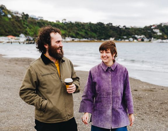Two people walking along a pebbled beach near the shoreline, with calm water, boats, and a tree-covered hillside in the background. One person is holding a takeaway coffee cup.