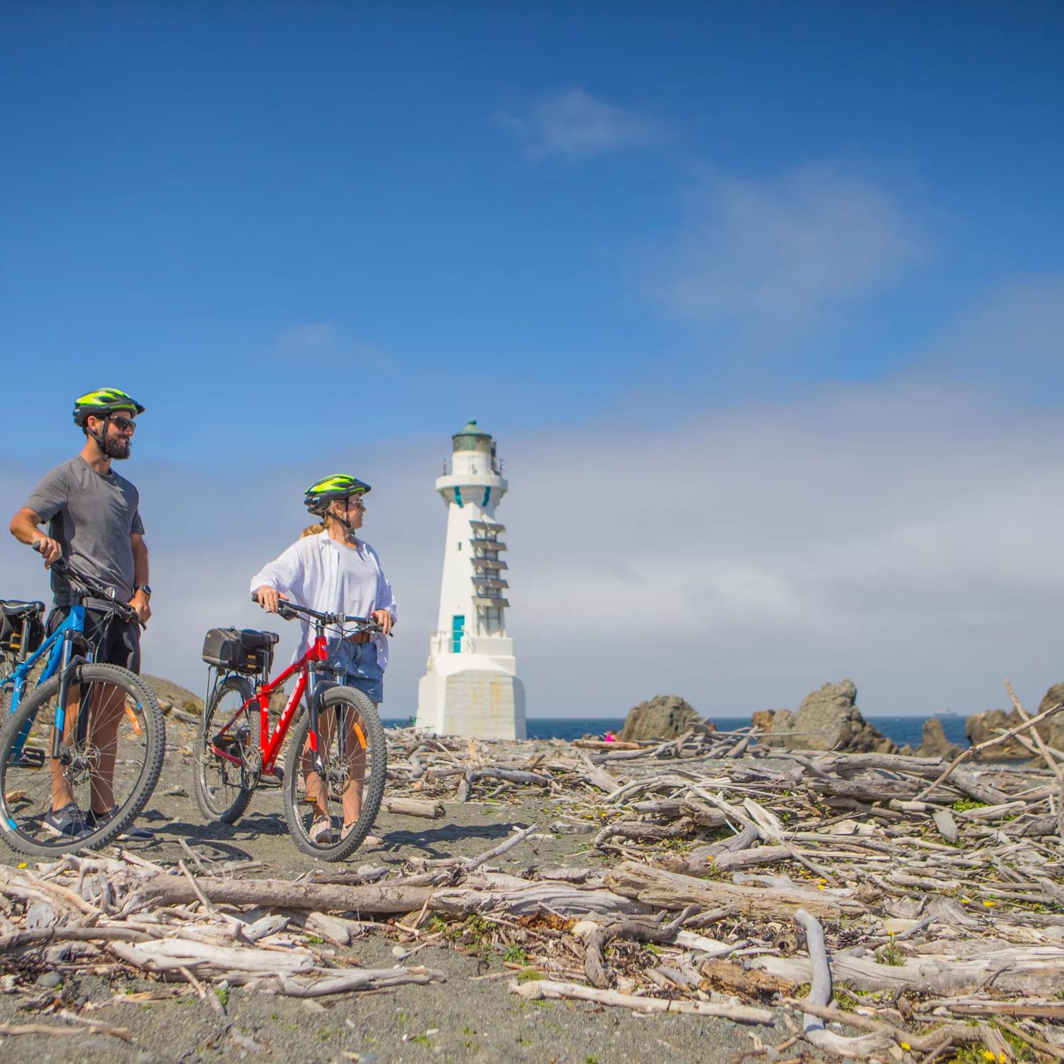 Two people stand with their bicycles on a beach with heaps of driftwood at Pencarrow Coast Road, a trail that starts from Burdan's Gate just south of Eastbourne. A small white lighthouse is seen in the background.