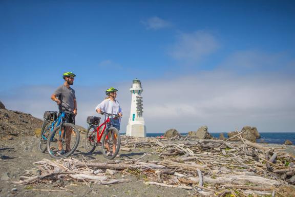 Two people stand with their bicycles on a beach with heaps of driftwood at Pencarrow Coast Road, a trail that starts from Burdan's Gate just south of Eastbourne. A small white lighthouse is seen in the background.
