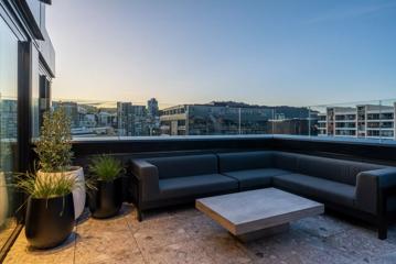 An urban balcony with a city view. There are three potted plants, a black sectional couch and a square cement table.