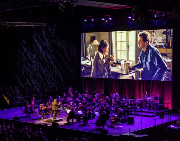 A full symphony orchestra plays the soundtrack from the film Love Actually on the stage at the Micheal Fowler Centre in Wellington with purple flood lighting and a scene from the film projected in the background. 