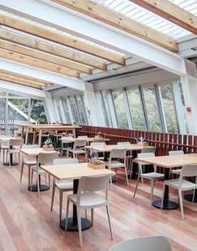 Inside a sheltered patio area of Zealandia, where tables and chairs are set up for a dinner setting. Views of native bush can be seen through the outside glass.