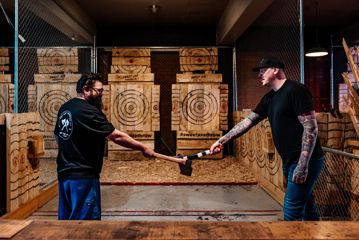 Two people reach out with axes before throwing towards wooden targets set up in front of them.