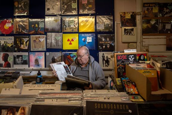 A man sits behind the counter looking through a stack of vinyls at Lo-Cost / Moonhop, a music store in Petone, Lower Hutt, New Zealand.